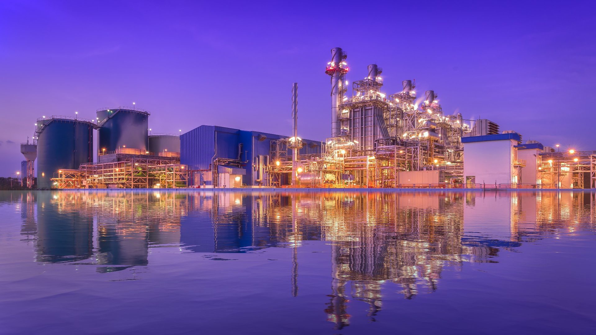 Industrial facility illuminated at night, reflecting on a calm body of water.
